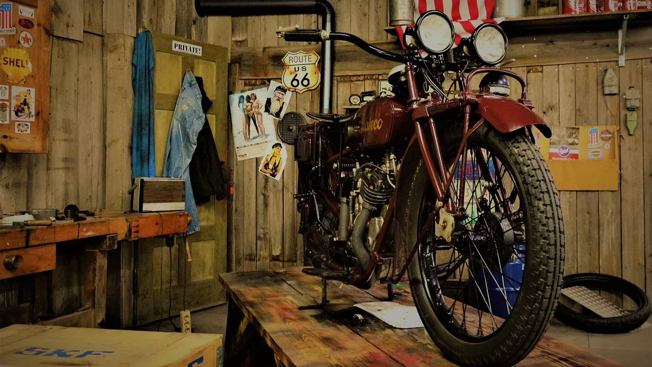 Najbardziej awaryjne motocykle sprzedawane w Ameryce 2 This classic-style motorcycle is parked on a wooden worktable in a vintage American garage. The background features authentic details like wood paneling, Route 66 and Shell oil signs, old tools, a radio, and classic posters. The bike is dark red with a large front tire and twin headlights, capturing a piece of American motorcycling history.