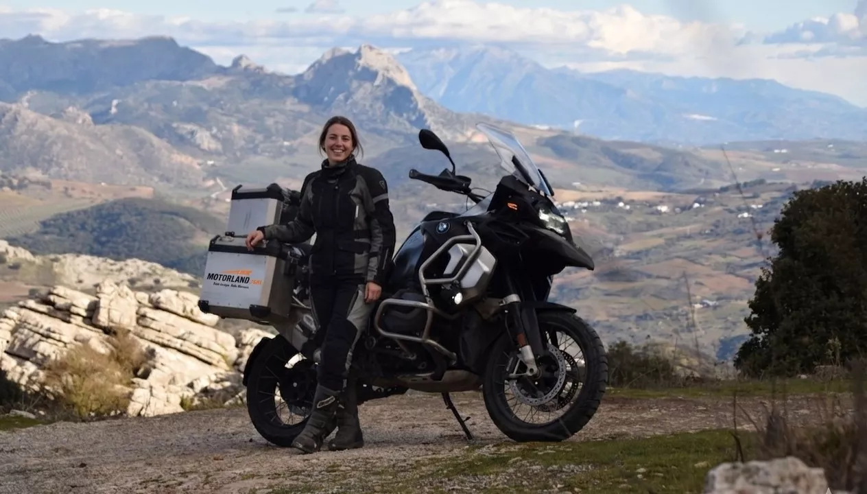 Młoda kobieta w stroju motocyklowym uśmiecha się przy motocyklu BMW GS z logo Motorland.rent na tle gór Andaluzji w Hiszpanii.